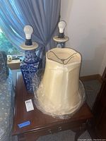 Two blue and white ceramic lamps on a wooden table with one lamp shade wrapped in plastic placed in front. Both lamps have bulbs installed but the shades do not fit on the lamps.