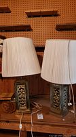 Pair of matching table lamps with ornate metallic bases and white pleated shades placed on a wooden surface against a pegboard background.