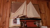 Model sailboat with black and red hull, mounted on wooden stand, white fabric sails fully rigged, garage background