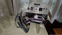 Shark Ultimate Professional iron G1568C in front of original box and small water measuring cup, placed on a table, near window with curtains.