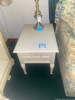 Single painted white side table with two drawers, turned legs, placed next to floral sofa.