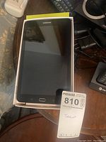 Black Samsung Galaxy Tab E tablet placed inside original white box on a round table, with Verizon green box underneath. Tablet screen is visible with Samsung logo and home button.