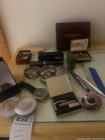 Full lot showing wristwatches, grooming kit, cuff links, locket, and trinket box on glass table.