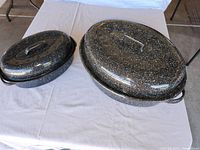 Two enamel-coated speckled roaster pans with lids on a table, showing size difference and condition