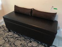 Front angled view of black faux leather bench with two brown lumbar cushions on a patterned carpet, showing surface texture and leg condition.