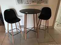 Bar height round table with black laminate top and natural wood legs with two black faux leather bar stools with wood legs and metal footrests.