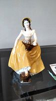 Front view of the Coalport 'Constance' porcelain figurine showing a woman in yellow dress sitting with basket.