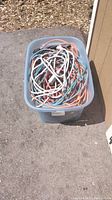 Plastic storage bin filled with assorted indoor and outdoor extension cords and a power bar outdoors on pavement near building.