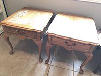 Front view of two wooden end tables placed side by side showing table tops and drawer fronts with metal pulls.