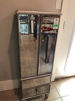 Front view of the mirrored jewellery cabinet showing two mirrored doors and two mirrored drawers at the base. Cabinet stands on four legs and has a silver mirrored finish with small damages.