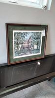 Framed artist's proof print leaning against a wall above a black surface, showing full framed artwork and part of window light.