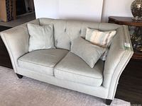 Front view of light green upholstered loveseat with tufted backrest, seat cushions, pillows, and visible nailhead trim.