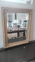 Full view of rectangular wall mirror with beveled glass and ornate gold-tone frame on black surface.