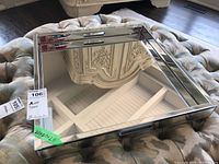 Square mirror tray with bevelled, double-layer mirrored edges, showing clear reflections of surrounding furniture, placed on a cushioned tufted ottoman.