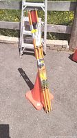 Two orange safety cones and six bundled orange safety markers leaning against a ladder, all placed outdoors on pavement.