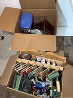 Open cardboard boxes showing contents including multiple spools of thread in a wooden rack and other items inside resulting boxes.