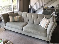 Front angled view of light greenish-gray upholstered sofa with 2 loose seat cushions, 3 reversible throw pillows, nailhead trim, and wood veneer legs in living room setting.