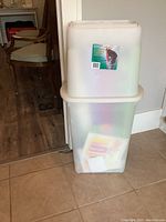 Two stacked translucent white plastic containers, upper container labeled for gift wrap storage, lower container filled with multiple rolls of holiday wrapping paper.