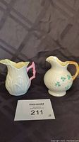 Both Belleek creamers (Lily White and Classic Clover) side by side on a dark gray patterned background.