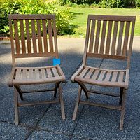 Front view of two teak folding chairs with slatted backs and seats on outdoor paved surface.