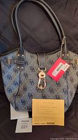 Front view of blue denim-style logo satchel with black leather trim, gold-tone logo lock clasp, two handles, and included yellow registration card and MaxSold lot tag.