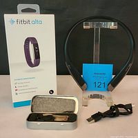 Fitbit Alta box standing upright next to LG wireless headset on display stand with charging cable and two additional wristbands in a metal case.