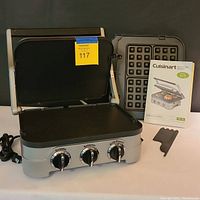 Open electric grill with reversible grill plates installed, extra waffle plates and instruction booklet visible beside unit.