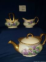 Front angle view showing teapot with lid and two other pieces (sugar bowl and creamer) on dark fabric background, highlighting hand painted African violet flowers and gold trim.