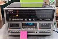 Front view of Soundesign stereo with dual cassette decks, control knobs, and digital clock display on silver panel with dust cover closed.