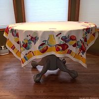 Full view of the rectangular tablecloth draped over a round table showing the white background and colorful Southwest-style border with pottery and gourds in yellow, red, blue, and aqua hues.