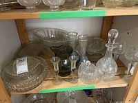 Shelf of assorted clear glassware including decanter with stopper, stemmed glasses, bowls, large round dish with lid, and ribbed pitcher. Items appear used and dusty.