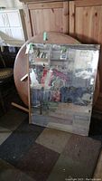Full front view of rectangular mirror leaning against wall, showing wood frame, reflective surface, and surrounding objects reflected.