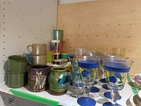 Overview of assorted mugs and cups including green ceramic mugs, purple decorative mug, ceramic pitcher, and clear floral glasses.