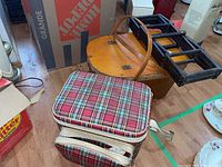 Photo of picnic baskets and wine rack on floor showing plaid cooler bag, folded wine rack, and woven basket with broken handle