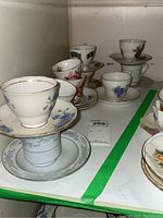 Photo of assorted vintage ceramic tea cups and saucers with floral designs placed on a shelf. Items show wear including chips.