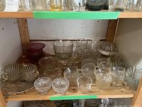 Shelf with various glass tea cups, small bowls, dishes, and a ribbed glass juicer. Items mostly clear glass with one maroon bowl, some with decorative edges and patterns.