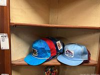 Overall view of seven men's ball caps stacked on a wooden shelf, showing various colors and logos.