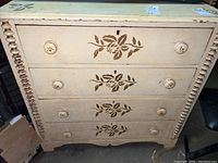 Front view of cream-painted wooden dresser with four drawers, each drawer featuring a brown floral applique design and round wooden knobs. Shows wear and chipping paint.