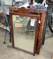 Two wood framed mirrors standing against a fence. One mirror is large with carved wooden top and finials, rich wood grain visible. Second mirror slightly smaller and plainer.