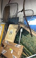 Photo shows two long handle catching nets resting against wall with paintings and boxed artwork in background.