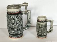 Pair of vintage ceramic beer steins with steam locomotive train designs, one with hinged lid, shown side by side on white background.