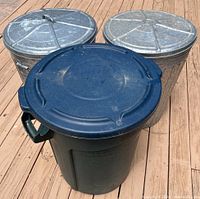 Photo of three trash cans on wooden deck; two galvanized steel with lids, one black plastic with lid on top, showing handles and worn condition.