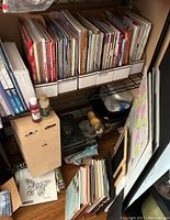 Shelf with multiple paperback books organized in white labeled boxes and some art supplies on the shelf and floor.
