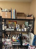 Wide view of three black metal shelves with assorted art supplies including paint bottles, brushes, art books, and craft materials, shelving unit not included.