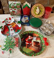 Photo of various Christmas-themed decorative plates, trays, tins, mini muffin pan, and fabric house decoration arranged on a table.