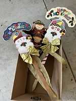 Four Christmas yard signs including two with snowmen, one Happy Holidays sign, and an elf figurine, arranged inside a cardboard box on concrete floor.