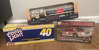Photo showing three boxed Coors collectible model vehicles: Ertl tractor trailer bank, Sterling Marlin transporter, and Monogram racing car kit.
