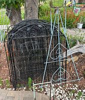 Nine black metal decorative garden fence panels stacked against a tree with a blue trellis in background