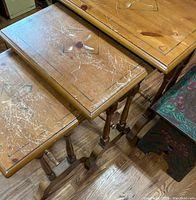 Three wooden nesting tables stacked; tops with visible scratches and inlaid designs including a central floral or organic motif and corner hearts.