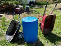 Blue plastic rain barrel centered with two wheelbarrows on each side in an outdoor garden area with grass and dirt.